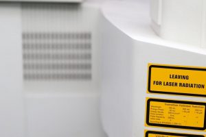 Gul varningsetikett för laserstrålning på en vit maskin med tekniska specifikationer.