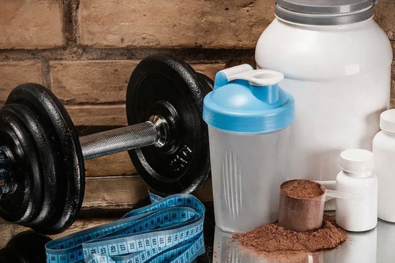 Hantel, shaker, måttband och kosttillskott framför en tegelvägg